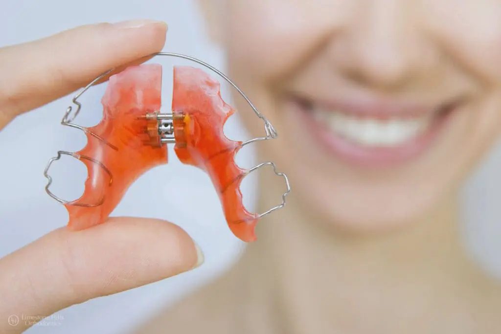 Smiling person holds a red, metal retainer close up at Limestone Hills Orthodontics in Austin, TX.