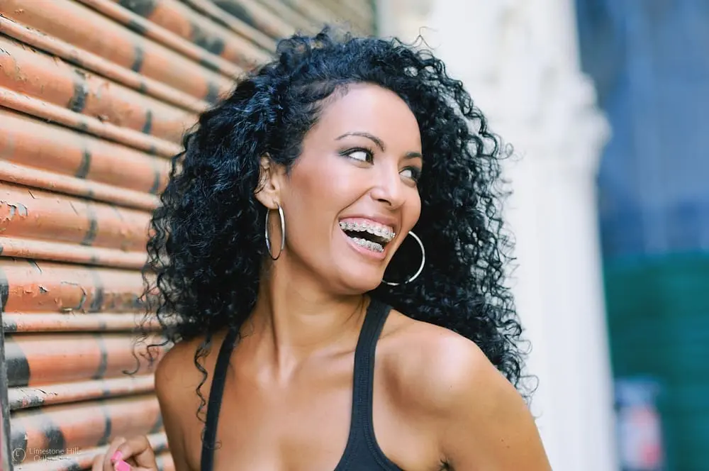 Adult woman with curly black hair and braces, wearing hoop earrings and a black top, smiling at Limestone Hills Orthodontics in Austin, TX.