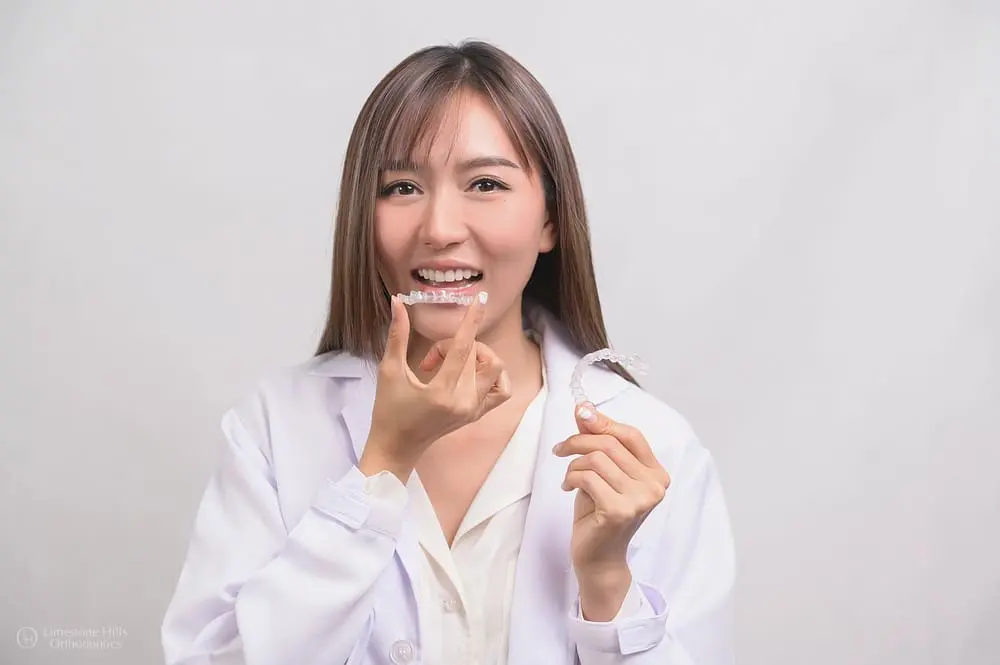 Smiling woman in a white coat holds Clear Aligners at Limestone Hills Orthodontics in Austin, TX, raising one toward her mouth.