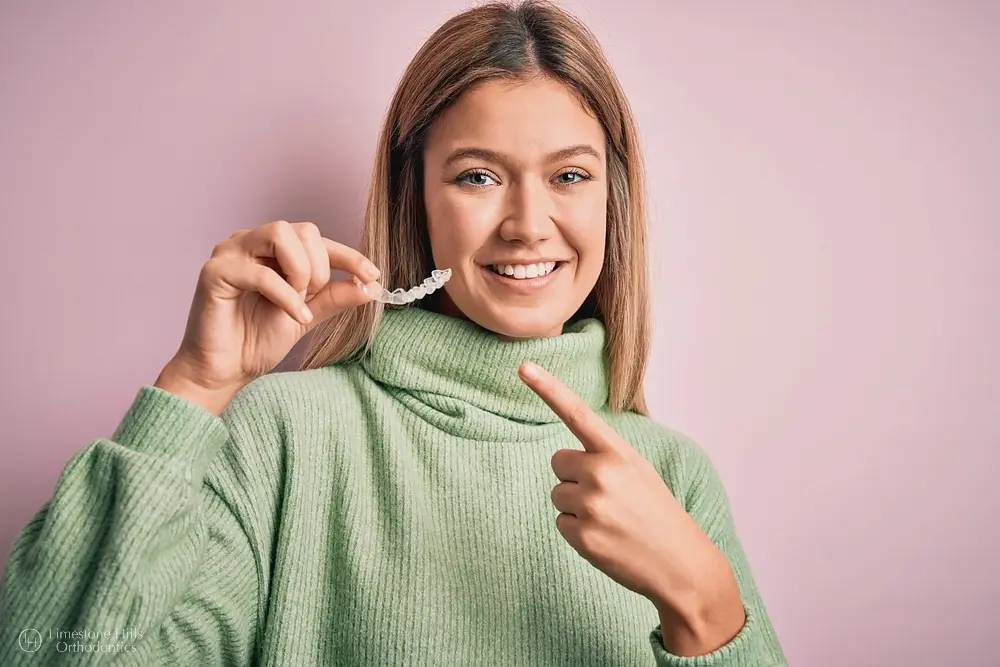 Smiling woman in green sweater points to aligner, referencing Limestone Hills Orthodontics in Austin, TX, and Invisalign insurance.